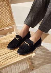 Zwarte suede loafers met franjes, gouden hardware en een ronde neus. Geplaatst op een geweven stoel met grijze maatpakken.
