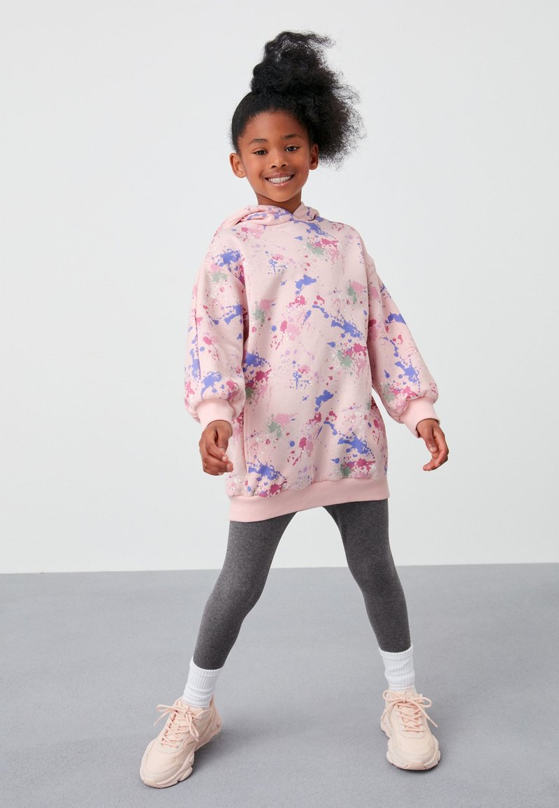 Ragazza in piedi con le gambe divaricate, indossa una felpa oversize rosa con stampa schizzi, leggings grigi, calzini bianchi e scarpe da ginnastica rosa, sorridendo verso la fotocamera.