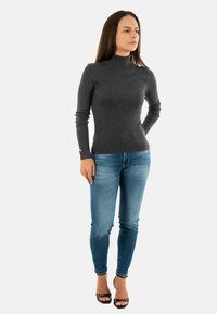 Maglione grigio a maniche lunghe con collo a imbuto e texture a coste, abbinato a jeans skinny blu e sandali neri con tacco alto.