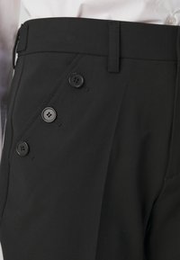 Pantalon noir en tissu à texture lisse, doté d'une poche inclinée et de quatre boutons noirs le long du côté avant.