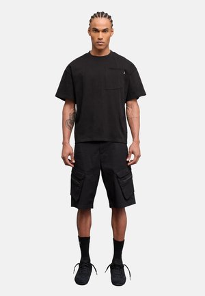 Hombre con cabello trenzado que lleva una camiseta negra de manga corta de gran tamaño, pantalones cortos cargo negros con bolsillos con cremallera, calcetines negros y zapatillas negras de pie recto.