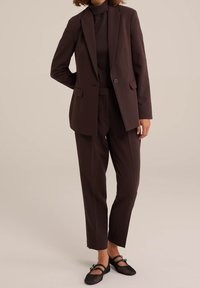 Kobieta ubrana w ciemno-brązową dopasowaną marynarkę i pasujące spodnie, w czarnych siateczkowych butach Mary Jane, na neutralnym tle.