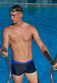 Nageur masculin portant un maillot de bain bleu foncé avec une accentuation bleue, peau mouillée brillant, s'agrippant à une échelle de piscine, devant une piscine bleue.