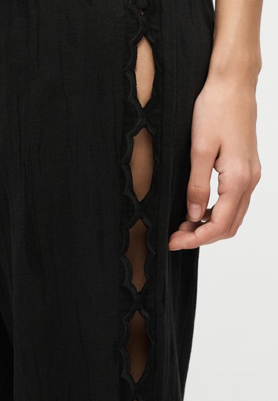 Κοντινό πλάνο ενός χεριού δίπλα σε μαύρο παντελόνι με κοψίματα με κυματιστό άκρο που αποκαλύπτουν το δέρμα κάτω από τη ραφή του ποδιού.