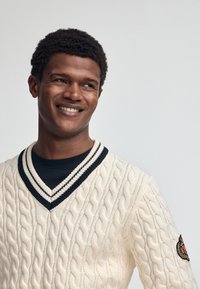 Kremowy sweter w warkoczowy splot z głębokim dekoltem w kształcie V, z granatowym i białym paskiem wykończeniowym. Zdobiony naszywką z logo na lewym ramieniu.