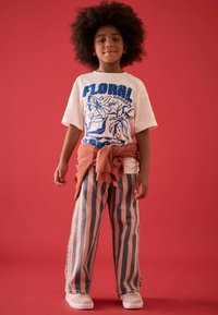 Camiseta blanca con estampado floral azul, suéter naranja atado a la cintura, pantalones de mezclilla a rayas rosas y azules, y zapatos blancos sobre un fondo rojo.