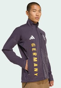 Fioletowa sportowa kurtka z wysokim kołnierzem, frontem na zamek, dużym żółtym napisem "GERMANY", kontrastującym teksturowanym wzorem po bokach oraz logo Adidas.