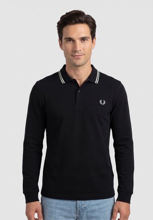 Uomo che indossa una polo nera a maniche lunghe con rifinitura bianca sul colletto e jeans azzurri, in piedi davanti a uno sfondo semplice.