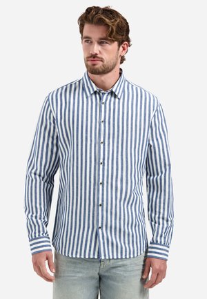 Mann trägt ein langärmliges Hemd mit blauen und weißen vertikalen Streifen und hellgraue Jeans, schaut leicht zur Seite.