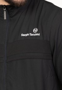 Schwarze Zip-Jacke aus glattem Stoff mit einem horizontalen Strukturband und dem weißen "Sergio Tacchini"-Logo im Brustbereich.