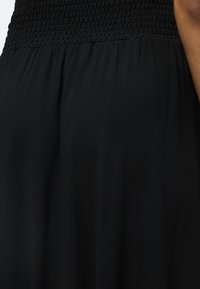 Tissu noir smocké qui passe à un tissu noir lisse sur un vêtement, avec une vue partielle du bras d'une personne sur le côté droit.
