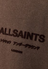 Tela marrón con superficie texturizada, que presenta el texto en relieve "ALLSAINTS" y letras adicionales en marrón oscuro.