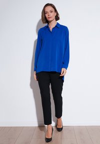 Blue long-sleeve shirt with a collar and button front, paired with black slim-fit trousers and black high-heeled shoes against a light backdrop.