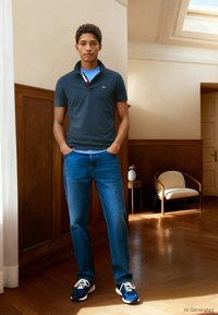 Marineblå polotrøye med en strukturert krage, kombinert med mellomblå jeans og marineblå joggesko med hvite lisser. En ensfarget bakgrunn med treinnslag.