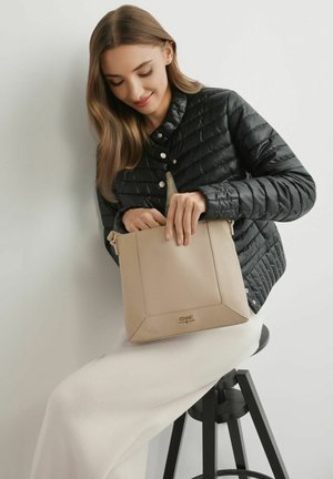 Kobieta ubrana w czarną pikowaną kurtkę i białą spódnicę siedzi na czarnym stołku, wkładając ręce do beżowej torebki na ramię.