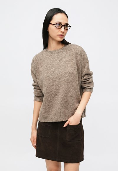 Femme portant des lunettes rondes à monture écaille de tortue, un pull en maille beige aux manches retroussées, et une jupe en daim brun foncé avec des poches à l'avant.