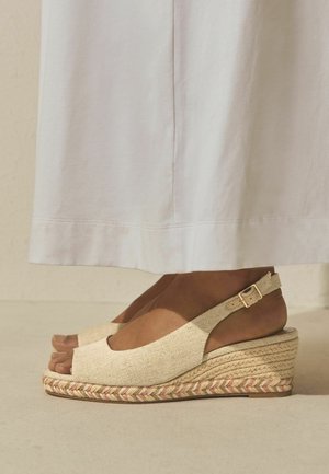 Feet wearing beige wedge espadrille sandals with ankle straps and open toes, paired with a long white skirt.