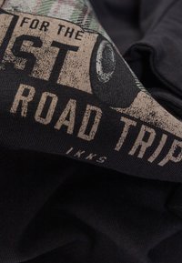Zwarte stoffen t-shirt met een vintage-stijl graphic met witte tekst en een geruite accent, die "ROAD TRIP" en andere details benadrukt.