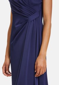 Marinblå klänning med mjukt tyg, diagonala veck och en tvistad design vid midjan. En närbild på en hand som rör vid klänningen.