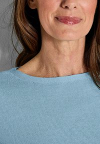 Hellblauer gestrickter Pullover mit weitem Ausschnitt, weicher Textur und subtilen Stoffmuster. Die Haare fallen sanft zur einen Seite.