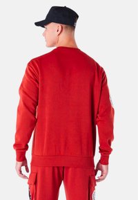 Felpa rossa con maniche lunghe, polsini e orlo a costine, con un fit casual e una texture morbida, abbinata a un cappellino nero.