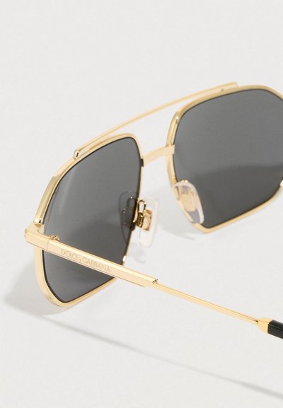 Γυαλιά ηλίου Dolce & Gabbana με χρυσή κορνίζα, σκούρους ορθογώνιους φακούς και ρυθμιζόμενα μαξιλαράκια μύτης σε ανοιχτό φόντο.