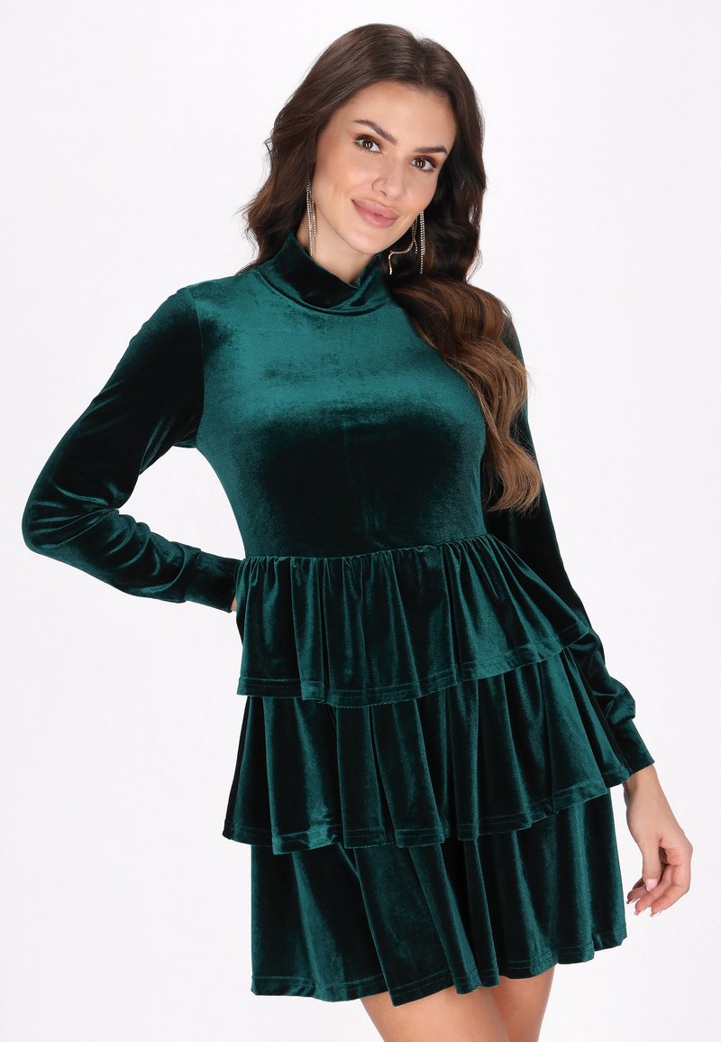 Rochie din catifea de culoare verde închis, cu guler înalt, mâneci lungi și volane stratificate la talie. Textură moale și detalii de design elegante.