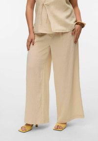 Un pantalon beige ample avec une finition texturée, associé à un haut assorti présentant un détail à nouer et des sandales à talons carrés jaunes.