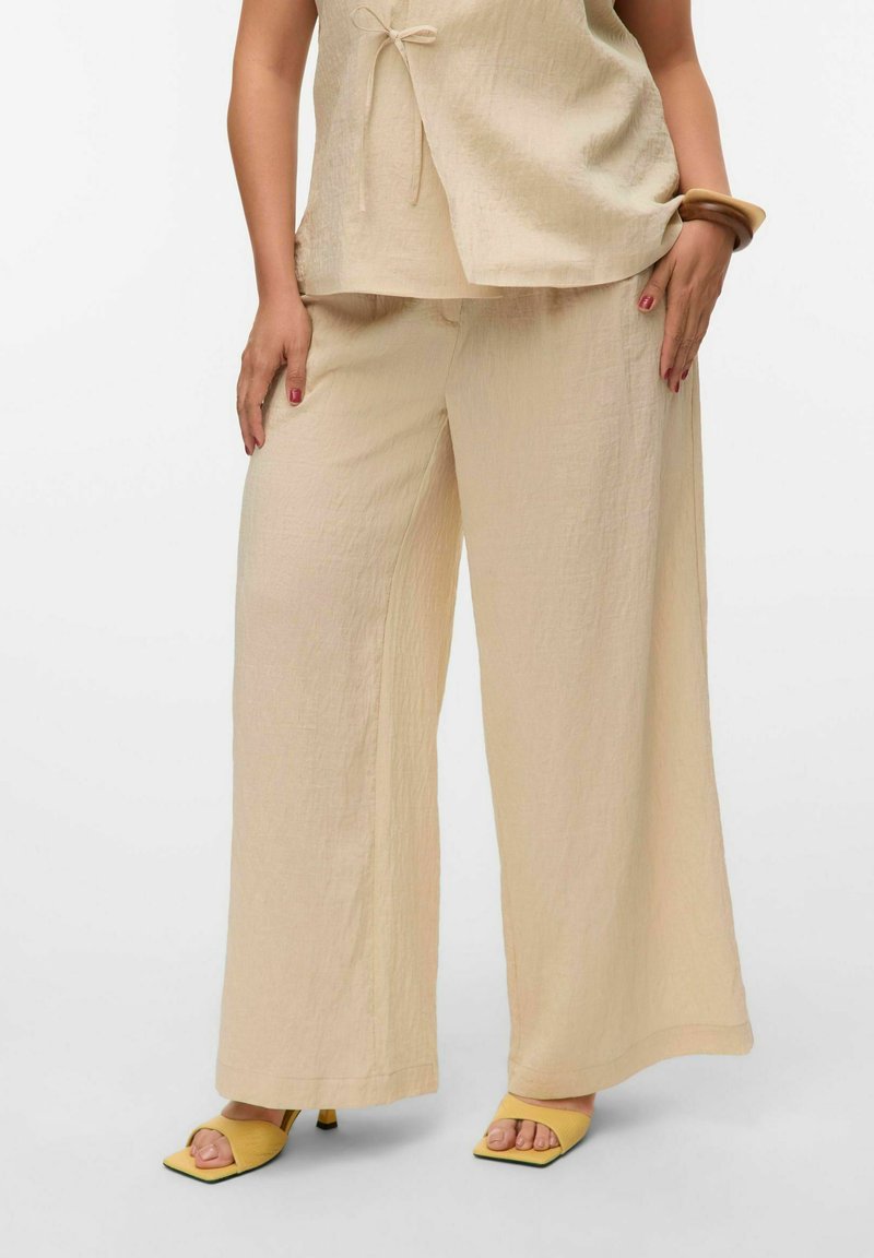 Un pantalon beige ample avec une finition texturée, associé à un haut assorti présentant un détail à nouer et des sandales à talons carrés jaunes.