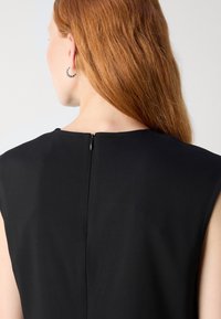 Femme aux longs cheveux roux portant un haut noir sans manches avec fermeture éclair dans le dos et une boucle d’oreille argentée en anneau, vue de dos sur fond blanc.