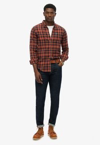Red and navy plaid shirt with button-down collar, paired with dark jeans and brown lace-up boots. Simple belt and watch accent outfit.