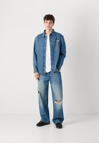 Camisa de mezclilla con botones y un bolsillo en el pecho, combinada con jeans anchos de tiro alto en azul claro desgastados y zapatos negros. Camisa blanca por debajo.