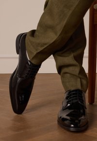 Zwarte gepatineerde leren dresschoenen met een puntige neus en intrikate veters, gecombineerd met lichtgroene linnen broeken, op een houten vloer.
