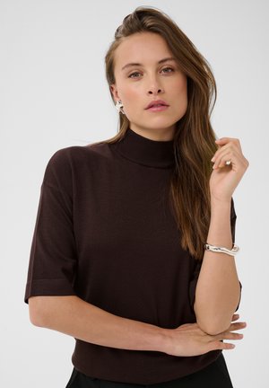 Femme aux longs cheveux bruns portant un col roulé à manches courtes marron foncé, des boucles d'oreilles argentées en forme d'étoile et un bracelet argenté à son poignet.