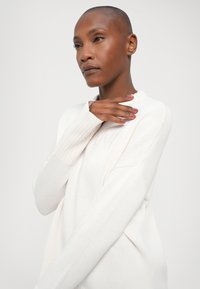 Pull en tricot blanc avec des poignets côtelés et une coupe décontractée. Présente un motif texturé et un col rond, mettant en avant un matériau doux et léger.
