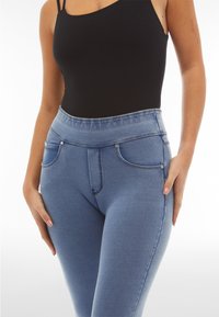 Nainen, joka pukeutunut korkeavyötäröisiin sinisiin farkkuihin ja mustaan hihattomaan toppiin, seisoo käsien rentona sivuilla.