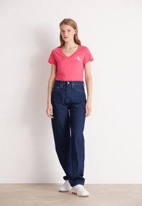 Camiseta de cuello en V rosa con logo, combinada con jeans de mezclilla azul oscuro de tiro alto y zapatillas blancas. Conjunto sencillo y casual.