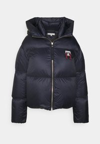 Marine blauwe pufferjack met een glanzende textuur, capuchon, voorzijde met rits en een rood-wit logo op de linkerborst. Verkorte ontwerp.