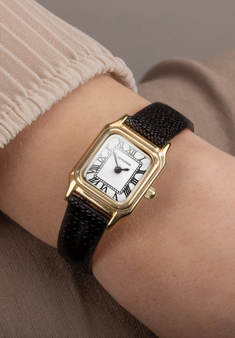 Goldfarbene quadratische Uhr mit einem weißen Zifferblatt, schwarzen römischen Ziffern und einem strukturierten schwarzen Lederarmband. Auf einem Handgelenk mit einem beigen Stoffärmel positioniert.