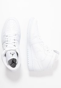 Hvite høyhælte sneakers laget av teksturert lær, med perforert tåområde, hvite lisser og en svart logo på tungen og hælen.