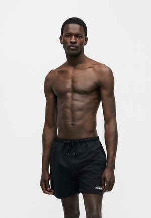 Μυώδης άνδρας που φοράει μαύρα μαγιό ellesse, στέκεται μπροστά σε απλό λευκό φόντο, κοιτάζοντας εμπρός με ουδέτερη έκφραση.