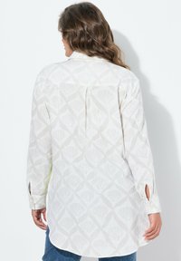 Lange mouwen shirt van lichtbeige stof met een subtiel schelp patroon, een rugplooitje en zijsplitten bij de manchetten.