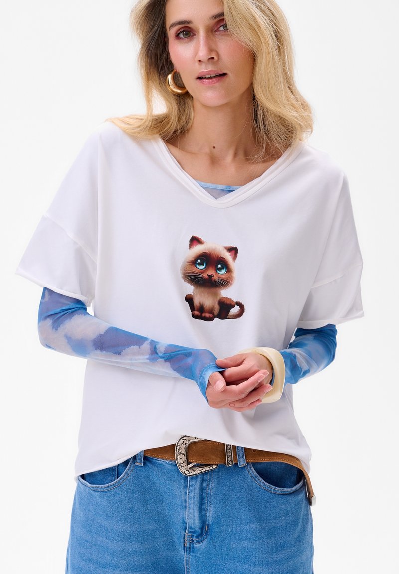 Biała koszulka z krótkim rękawem z grafiką kota w stylu kreskówkowym, pod spodem włożona jasnoniebieska koszula z długim rękawem w chmurki.