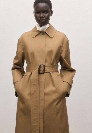 Mujer con abrigo trench camel con cinturón, gran cuello y bolsillos, manos en los bolsillos, de pie frente a un fondo liso y claro.