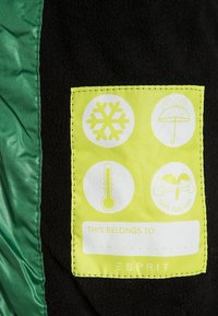 Černá tkanina se zeleným pruhem ukazuje obdélníkovou nálepku v neonově žluté barvě s bílými ikonami pro bezpečnost při počasí a vlastnictví.