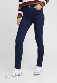 Jeans elásticos azul oscuro con un corte ajustado, cinco bolsillos y cierre con botón en la parte delantera, combinados con zapatillas blancas. Se ve una camiseta de mangas largas a rayas.