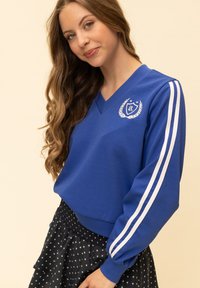 Blauwe sweatshirt met V-hals, witte strepen op de mouwen en een logo met lauwerkransontwerp. Het materiaal lijkt glad; gedragen met een zwarte rok met sterpatroon.