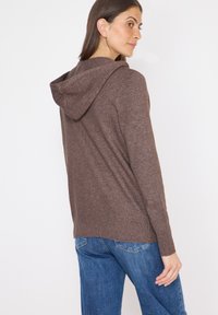 Brauner, strukturierter Kapuzenpullover mit geripptem Saum und Bündchen, kombiniert mit blauen Jeans. Sicht von hinten, die die Kapuze und den lockeren Schnitt zeigt.