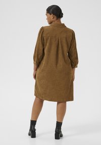 Woman with hair in bun wearing a brown corduroy knee-length dress with 3/4 puff sleeves and black heeled ankle boots, facing away.