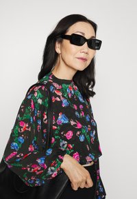 Blusa preta floral com padrões vibrantes em rosa, azul e verde; mangas longas; emparelhada com óculos de sol oversized pretos e uma bolsa preta.